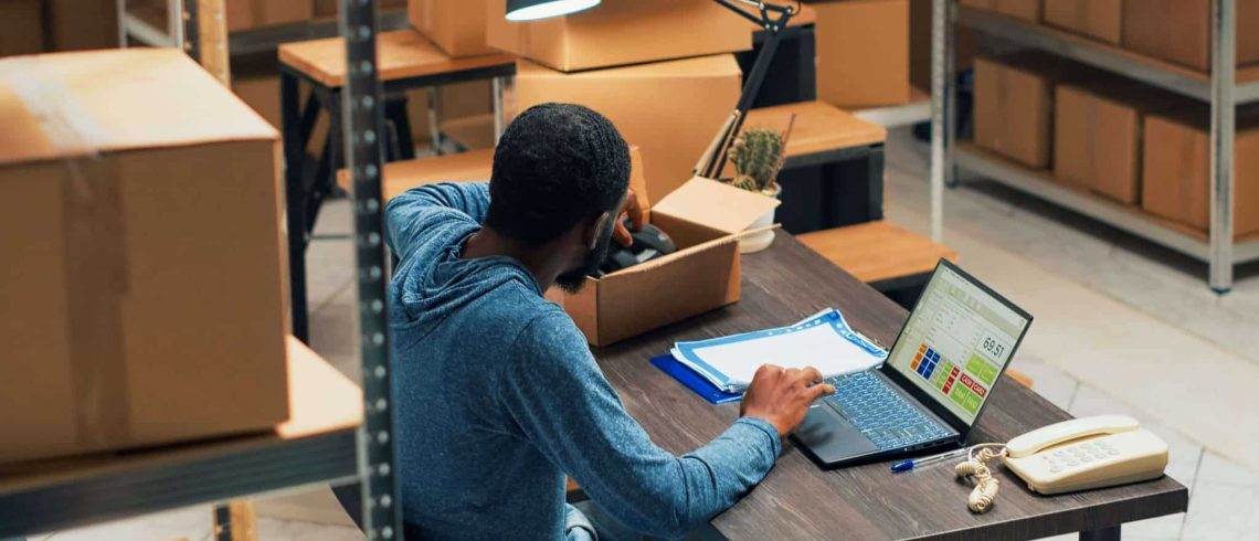 homem, sentado em uma mesa, na frente de um notebook, dentro de um estoque, analisando um produto na caix