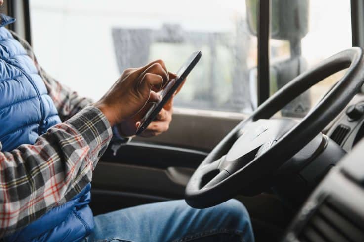Foto em close do interior da cabine de um caminhão, mostrando as mãos de um motorista com camisa xadrez e colete azul digitando em um smartphone próximo ao volante. A imagem ilustra o comportamento de risco e a distração ao volante frota.