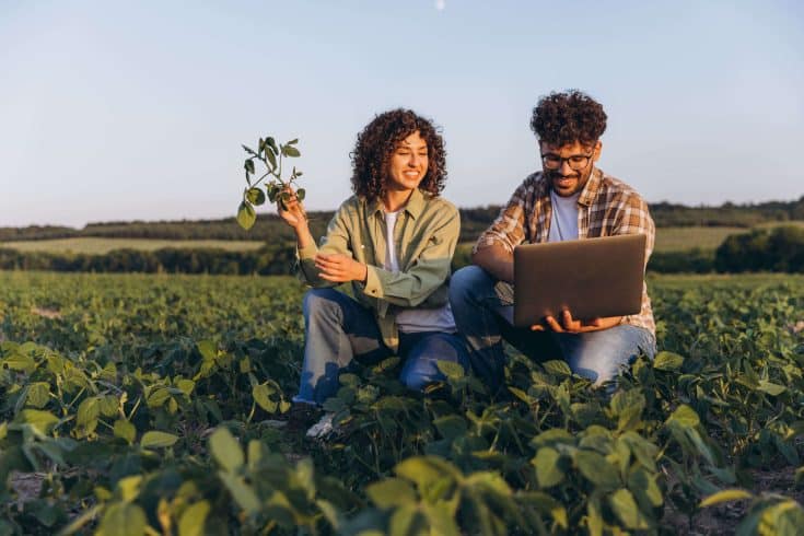 Dois produtores no campo analisando dados em um notebook na plantação. Representa a gestão tecnológica e a previsibilidade de entrega no agronegócio.