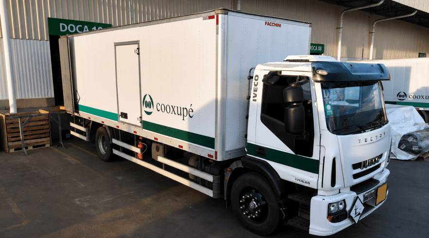 Caminhão baú Iveco Tector branco, com listra verde e o logotipo da Cooxupé na lateral e cabine, estacionado em frente à Doca 10 na área de carregamento de um centro de distribuição iluminado pela luz do dia.